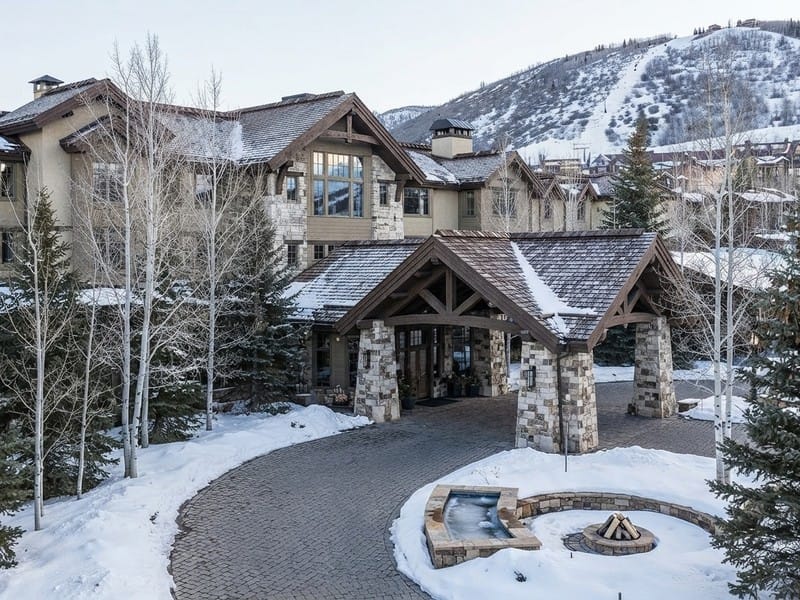 An exterior view of a stone and timber mountain lodge with a covered porte-cochere entrance, snow-covered grounds, a curved paved driveway, and ski slopes in the background.