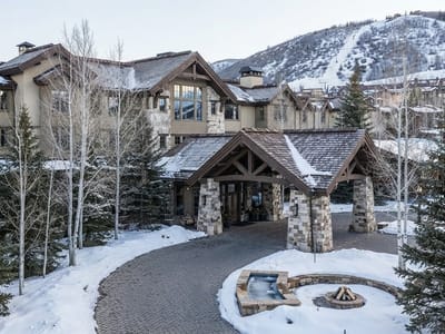 An exterior view of a stone and timber mountain lodge with a covered porte-cochere entrance, snow-covered grounds, a curved paved driveway, and ski slopes in the background.