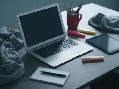 A wooden desk holding an open laptop, a tablet, highlighters, a notebook with a pen, a mug with pencils, and gray knit fabric.