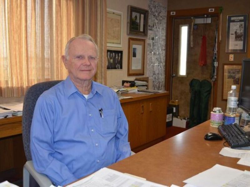 An older man in a blue dress shirt seated at his office desk surrounded by papers and framed photos